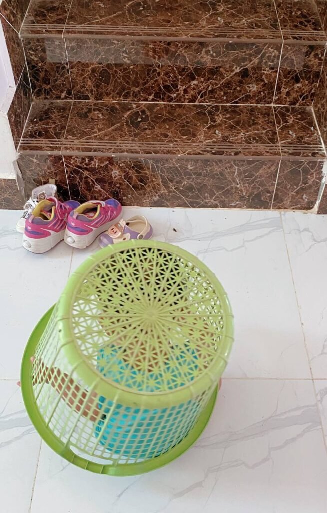 A toddler hiding underneath an upside-down green laundry basket on a white tiled floor next to pairs of children's shoes at the bottom of a staircase.