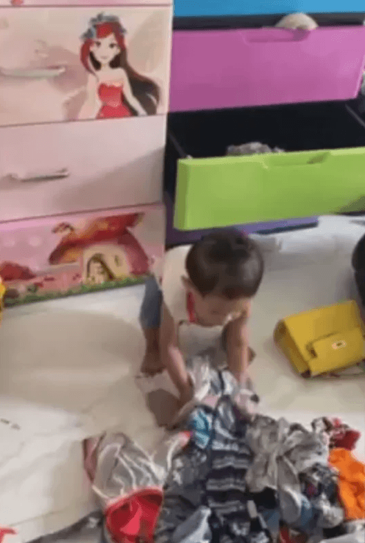 A young boy kneeling on the floor, playing with a scattered pile of colorful clothes next to his pink and green storage drawers.