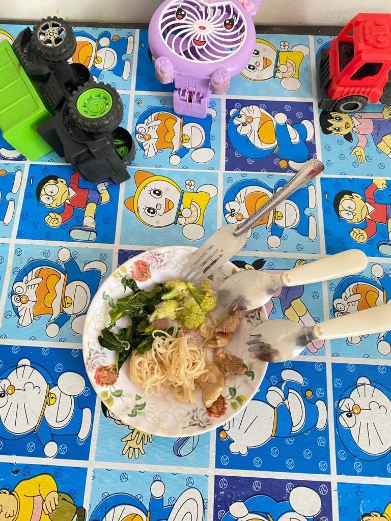Toddler plate with steamed broccoli, meat, and noodles next to a stainless steel fork and toy trucks.