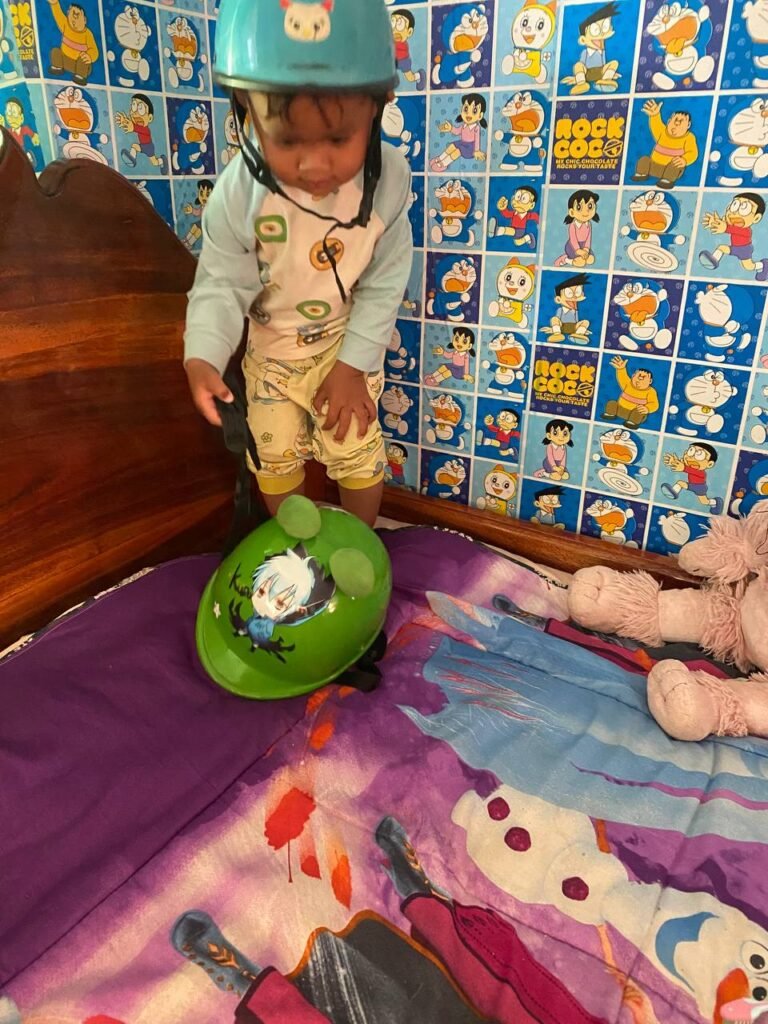 Toddler wearing a blue helmet looking down at a green toy helmet lying on a bed next to a pink stuffed poodle.