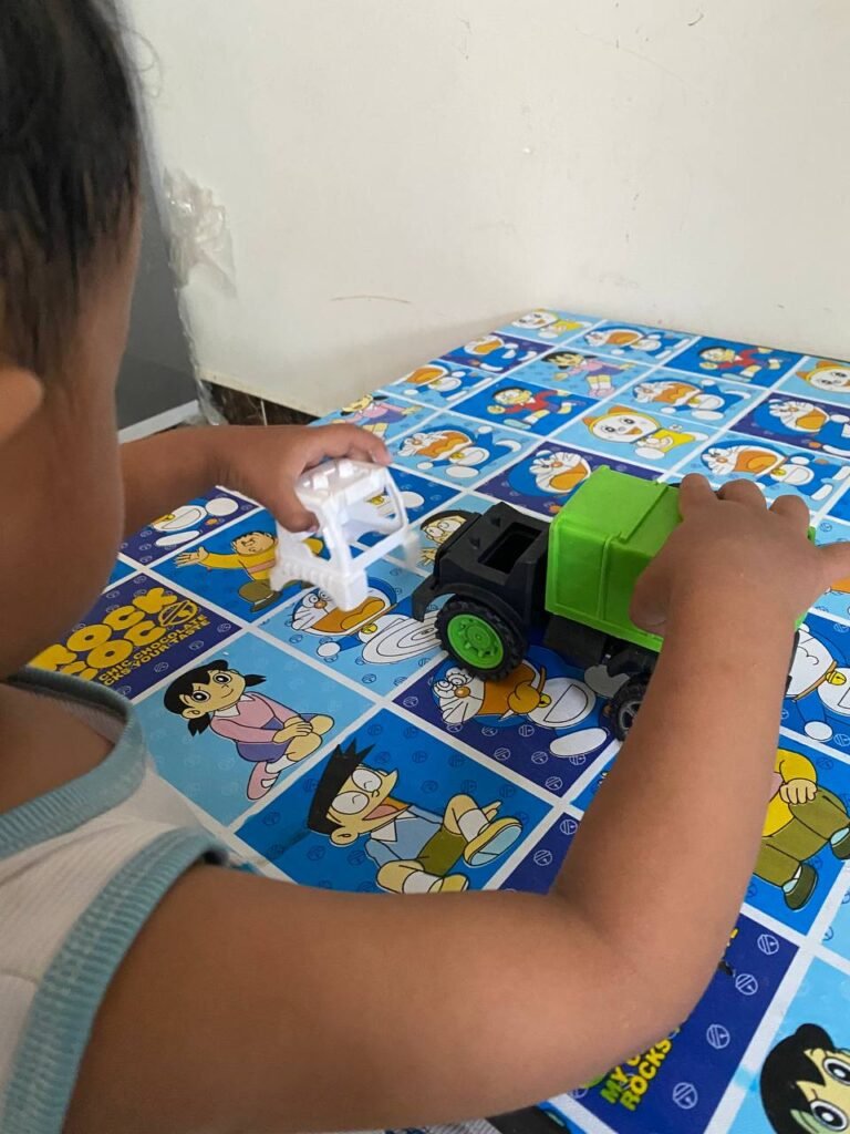A toddler boy holding a broken white piece of a green toy recycling truck, looking at the pieces.