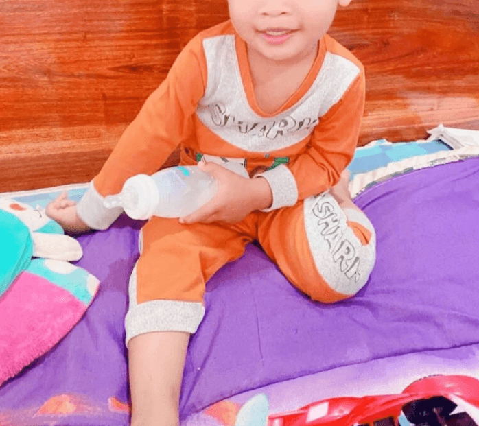 Toddler sitting peacefully on a purple mattress holding a milk bottle in his preferred sensory-friendly outfit.