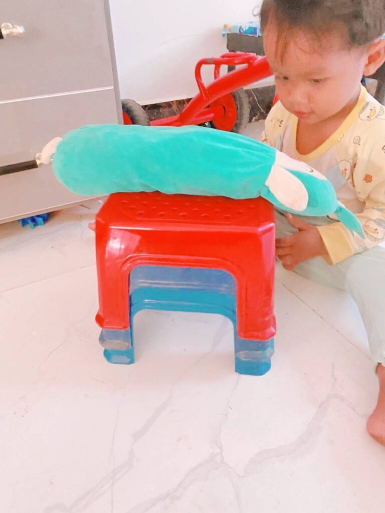 Toddler carefully balancing a long blue bolster pillow on top of a red plastic stool.