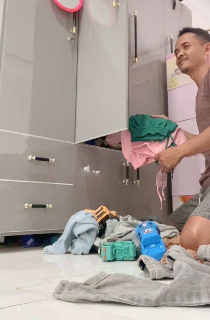 Father kneeling on the floor folding a pile of toddler clothes next to an open wardrobe.