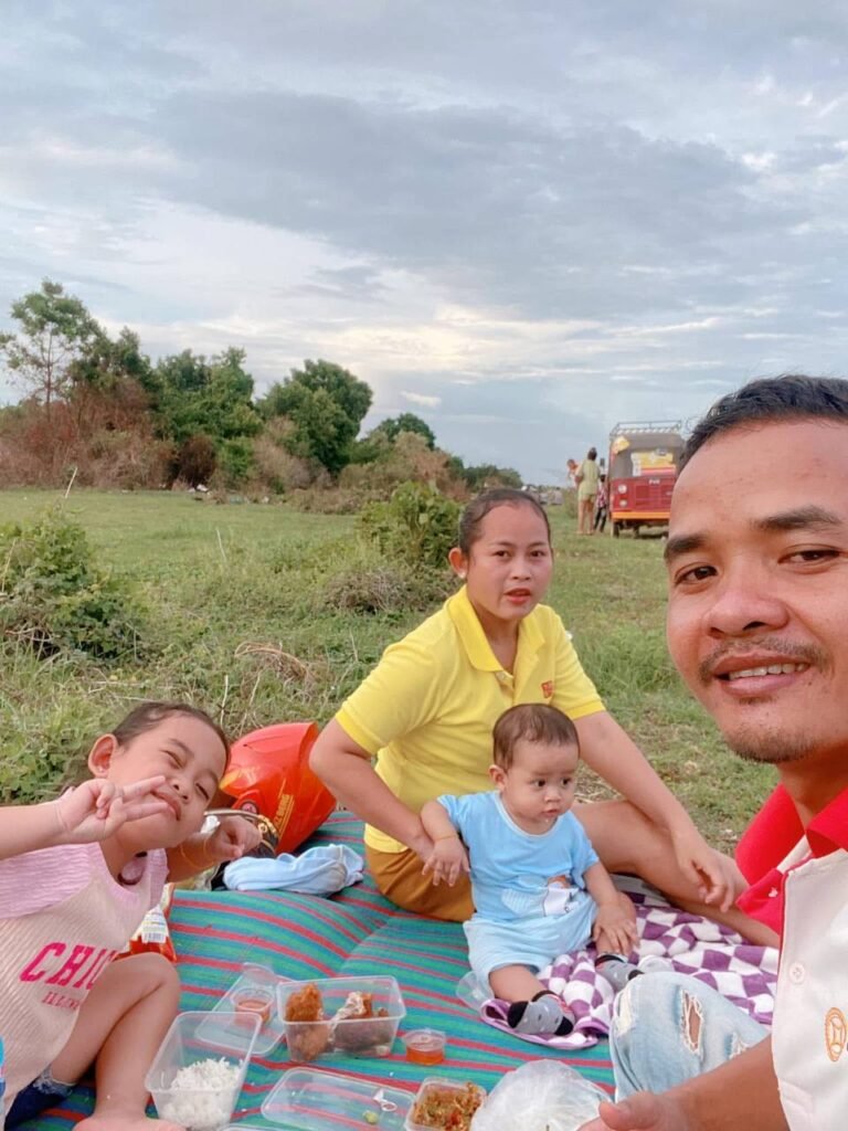 A family sitting on a mat in a grassy field eating rice and fried chicken directly from plastic storage containers.