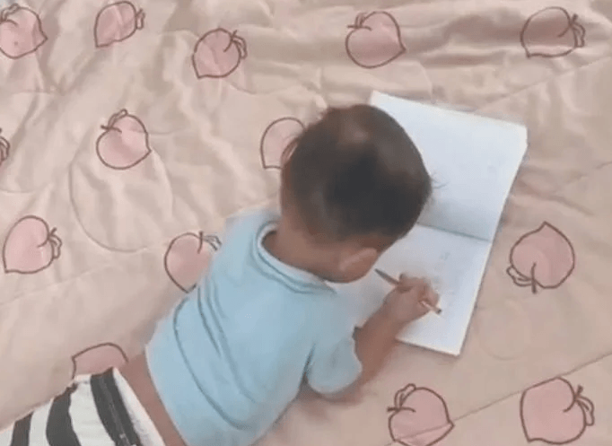 View from behind of a toddler lying on his stomach, deeply focused on scribbling in a blank notebook.