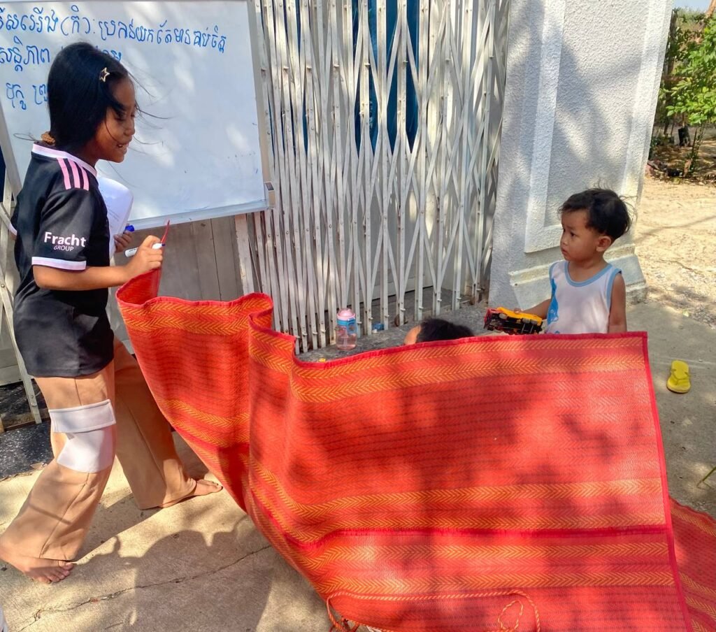 A gust of wind blows a red mat up over children playing school, causing the teacher and students to laugh.