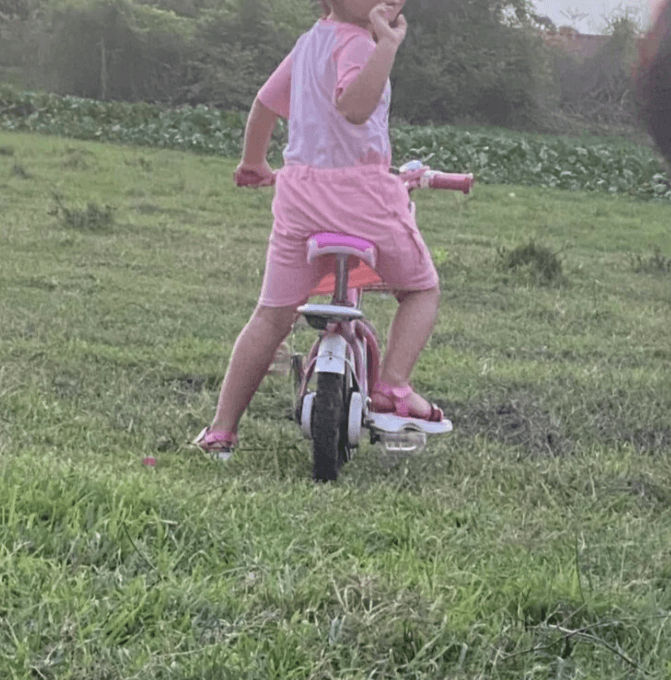 A young girl in a pink outfit rides her pink bicycle through a large, open grassy field.