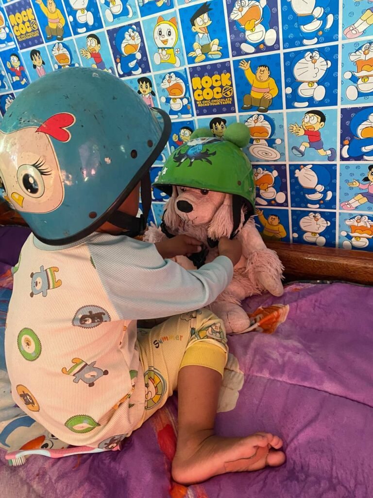 Close-up of toddler hands carefully buckling the chin strap of a green helmet onto a pink stuffed poodle toy.