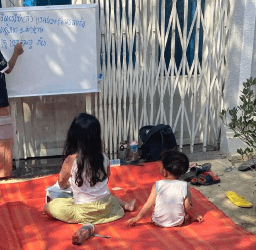 Three children sitting on a red woven mat in a driveway, playing school with a white board that has Khmer script written on it.