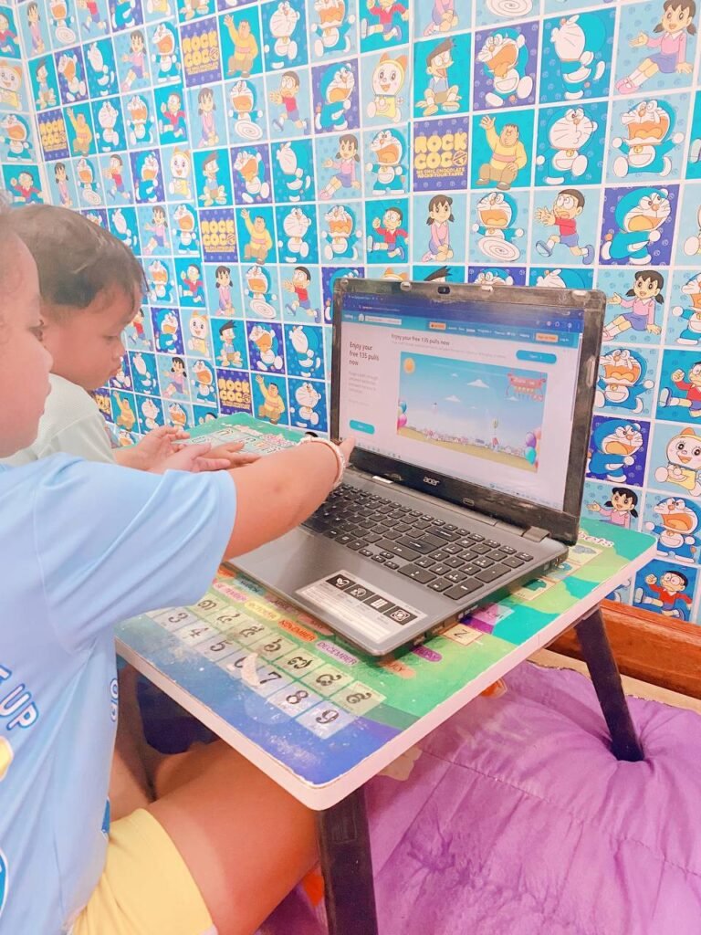 Little girl and toddler boy sitting at a low table playing the Typing Balloon educational browser game on a laptop.