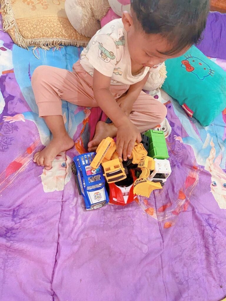 3-year-old child holding a group of different toy vehicles including a police car, construction truck, and garbage truck.
