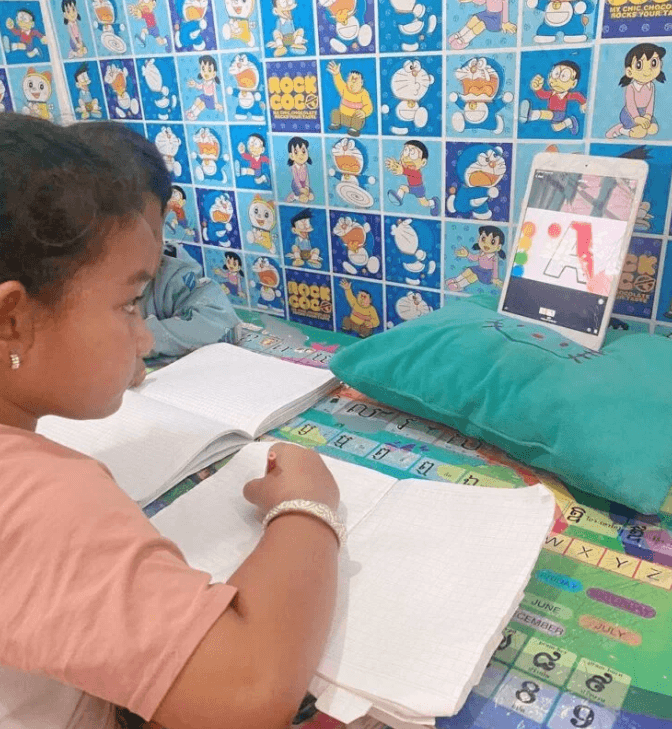 Older sister sitting at a green kid-sized desk in a Doraemon-themed room, using a tablet to practice writing the letter A.