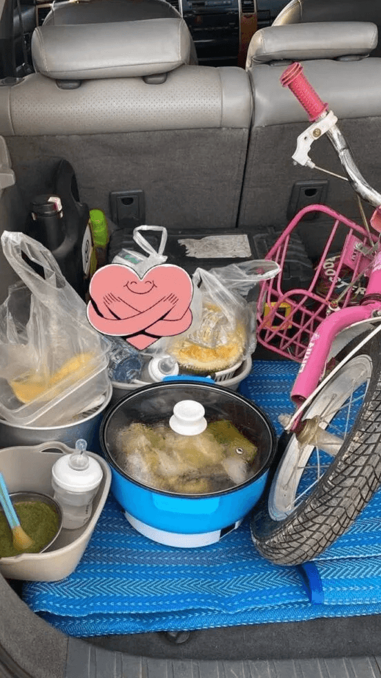 A car trunk packed for an impromptu family dinner picnic, featuring a blue electric cooker full of food right next to a child's pink bicycle.