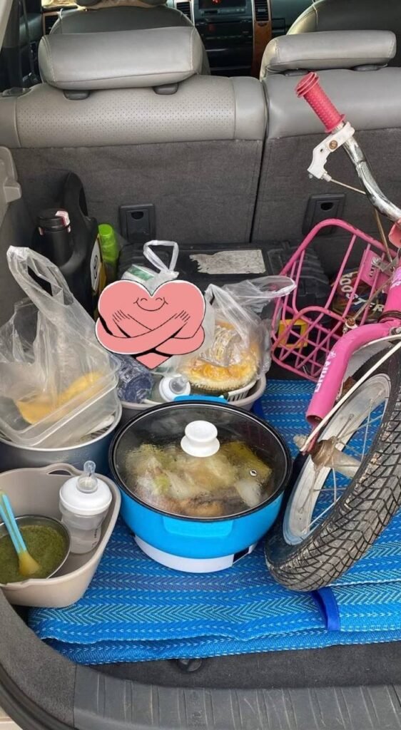 A car trunk packed with a blue electric cooker pot, groceries, and a pink child's bicycle for an outdoor dinner.
