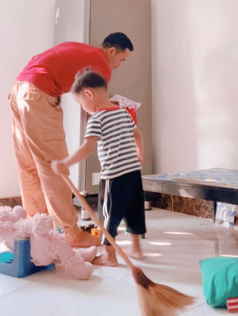 Father and toddler son sweeping the floor together amidst a messy room.