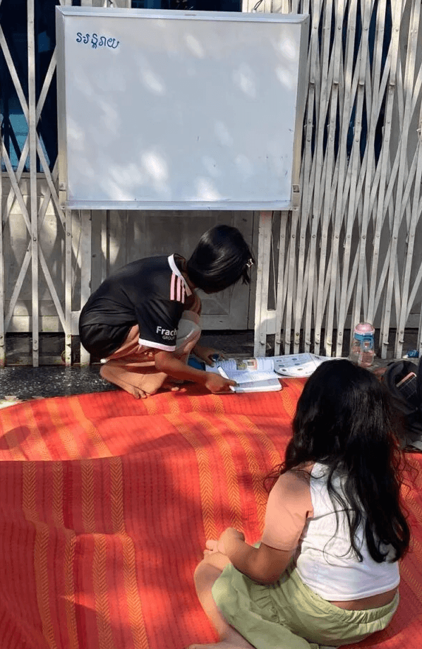An older girl acting as a teacher crouching down next to a younger student to look at a textbook together during pretend play.