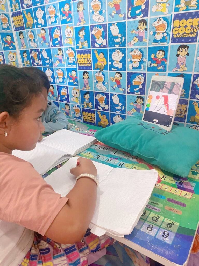 Toddler boy sitting next to older sister at a low green table with Doraemon wallpaper while she practices writing.