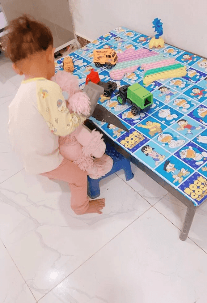 A toddler boy lifting a pink stuffed poodle to sit on a blue plastic stool next to his play table.