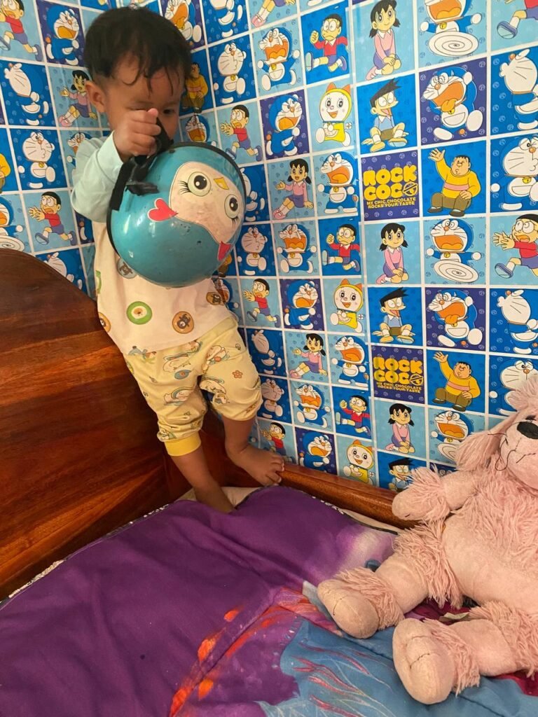 Toddler boy holding a blue cartoon bike helmet in a room with Doraemon wallpaper, getting ready for a ride.