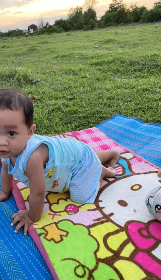 A toddler boy in a blue shirt sits on a colorful Hello Kitty picnic mat on the grass, exploring the outdoors.