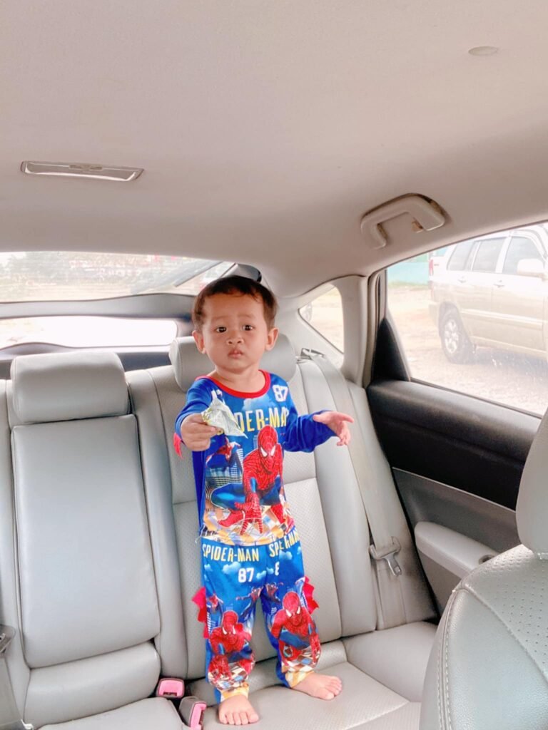 A two-year-old boy wearing blue Spider-Man pajamas standing seriously on the grey leather back seat of a car during a road trip.
