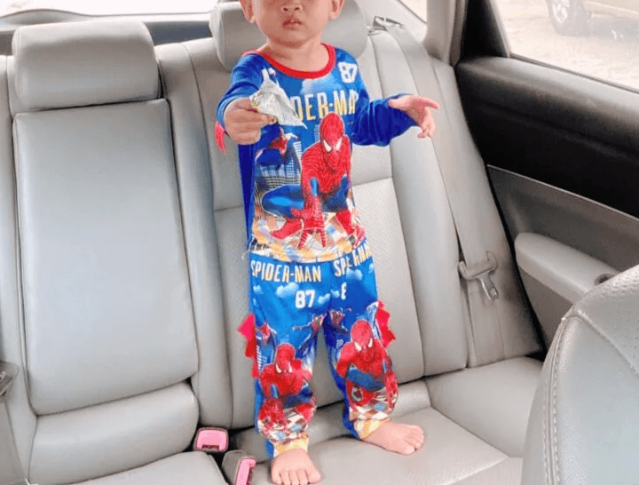 A two-year-old boy wearing blue Spider-Man pajamas standing seriously on the grey leather back seat of a car during a road trip.