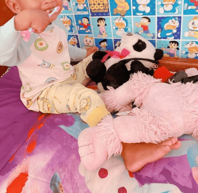 A toddler sitting next to stuffed animals happily brushing his own teeth with a pink toothbrush.