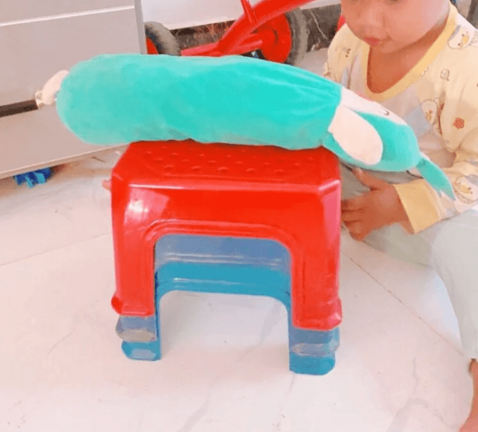 A creative structure built by a toddler using stacked red and blue plastic stools with a long blue pillow balanced on top.