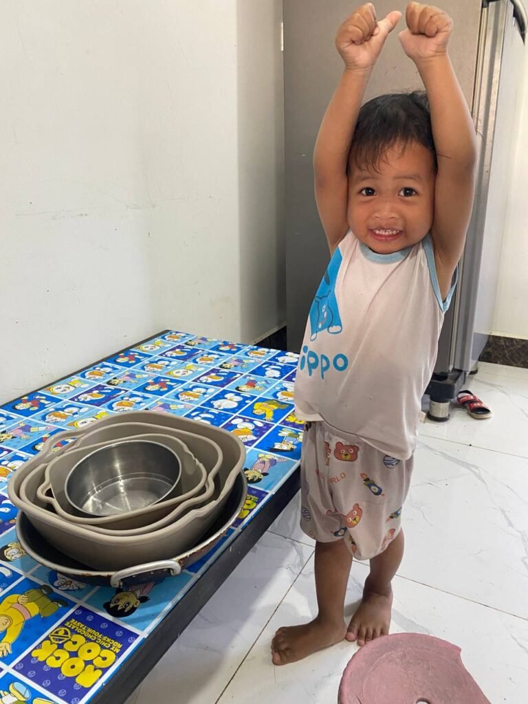 A happy toddler boy throwing his hands in the air to celebrate successfully stacking a tower of kitchen bowls.