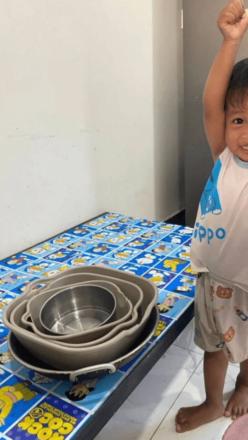 A happy toddler boy throwing his hands in the air to celebrate successfully stacking a tower of kitchen bowls.