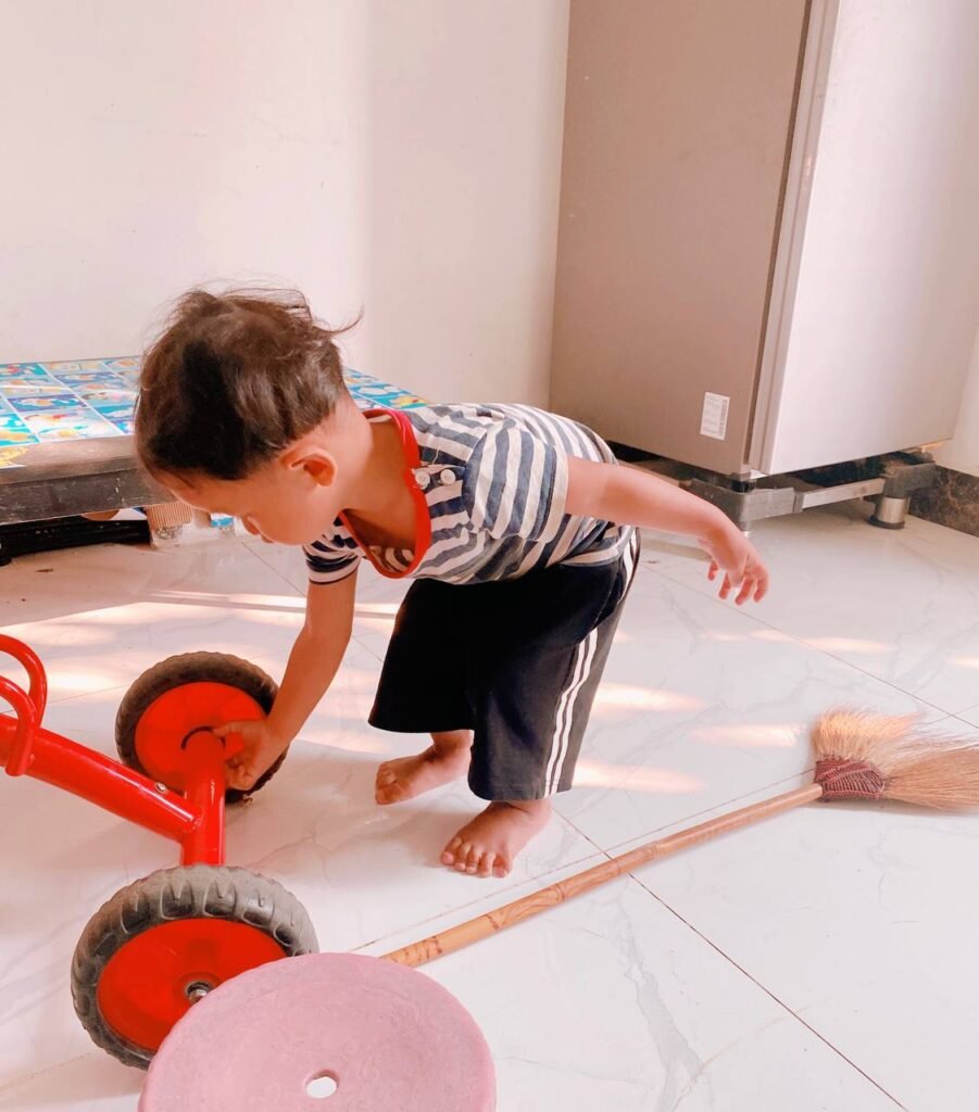 Two-year-old bending down to clean the wheel of his red tricycle with a broom.