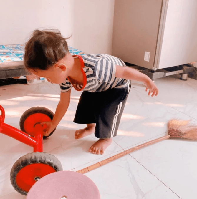 A toddler crouching down to inspect and clean the wheels of his red tricycle while doing chores.