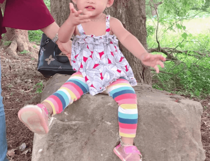 A three-year-old girl in colorful striped leggings sits on a rock at Ek Phnom Temple in Cambodia, holding up her hand while her mother gently steadies her.