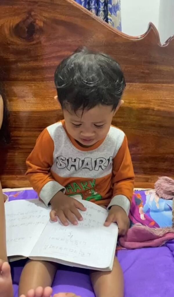 A 2-year-old boy wearing an orange shark shirt sitting on a bed and joining a family study session.