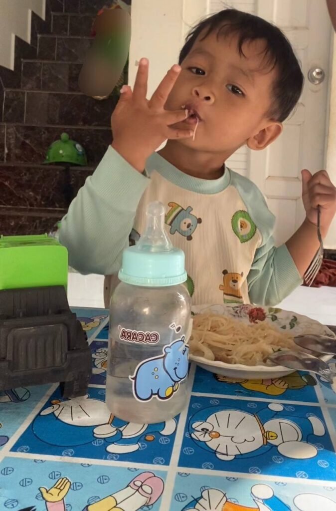 Toddler eating pasta with his fingers while holding a fork in the other hand.
