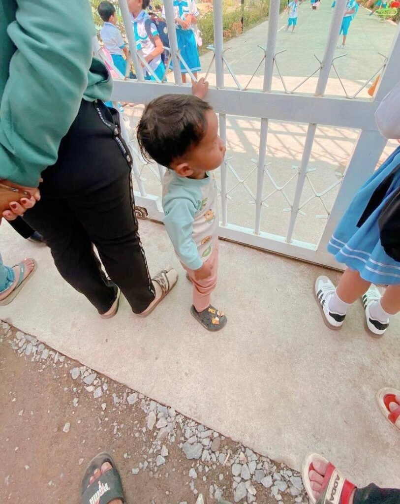 A toddler boy standing confidently at a school gate wearing black sandals with cartoon charms after overcoming shoe refusal phase.