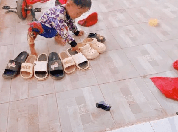 A two-year-old crouching down to carefully adjust a beige clog in a line of shoes, demonstrating fine motor control.
