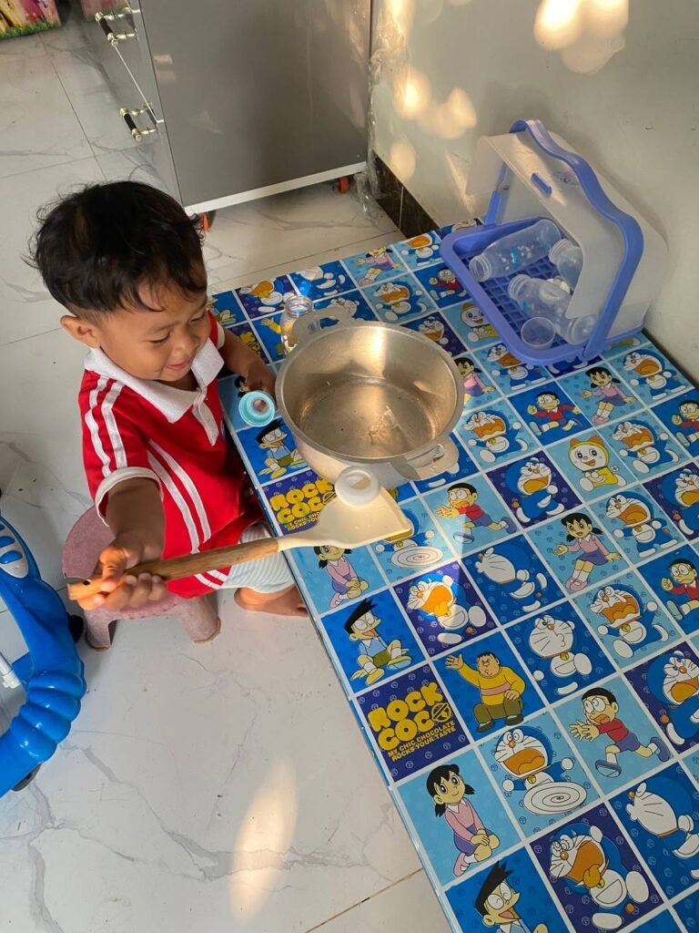 Toddler boy sitting at a table stirring empty milk bottles in an aluminum pot for a pretend play activity.