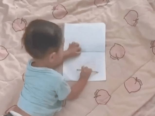 Top-down view of a toddler using a full fist grip to hold a pencil while scribbling in a notebook on the floor.
