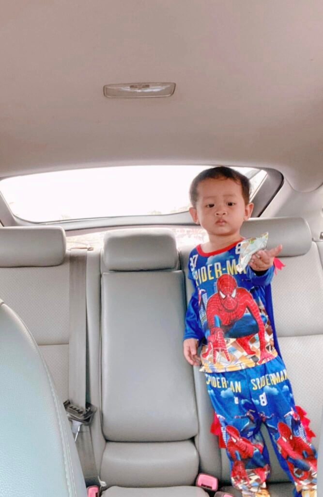 A young toddler in a Spider-Man suit handing an empty silver snack wrapper toward the camera from the back seat of a car.