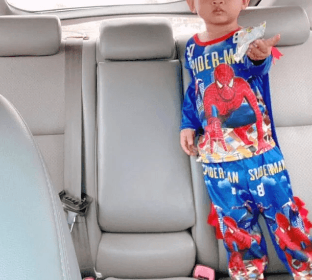 A young toddler in a Spider-Man suit handing an empty silver snack wrapper toward the camera from the back seat of a car.