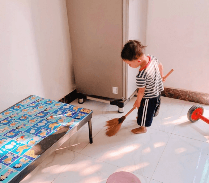A young boy using a broom to sweep the kitchen floor next to the refrigerator.