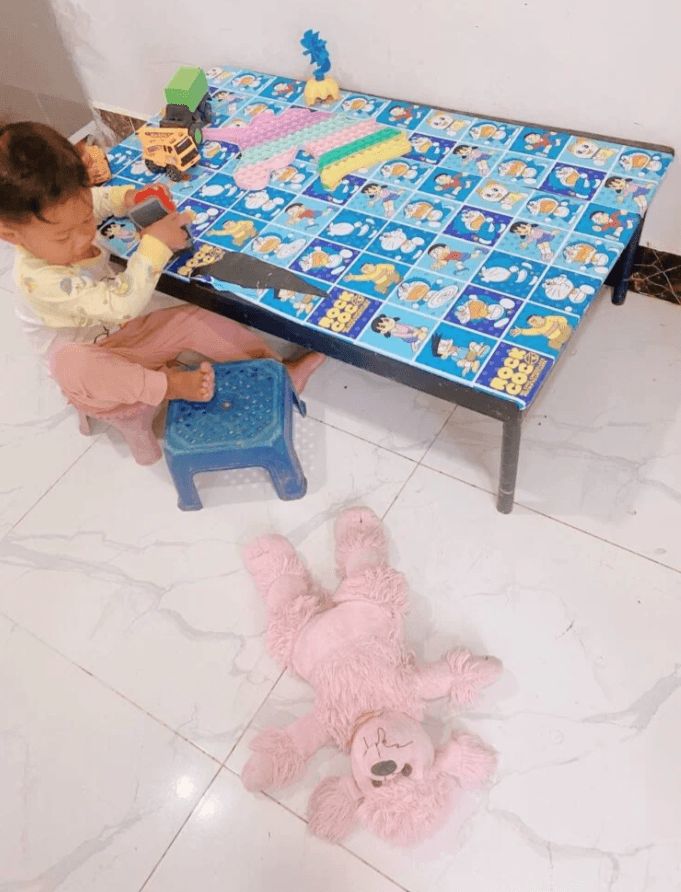 A toddler boy playing at a Doraemon table with toy trucks while a pink stuffed poodle lies forgotten on the floor.
