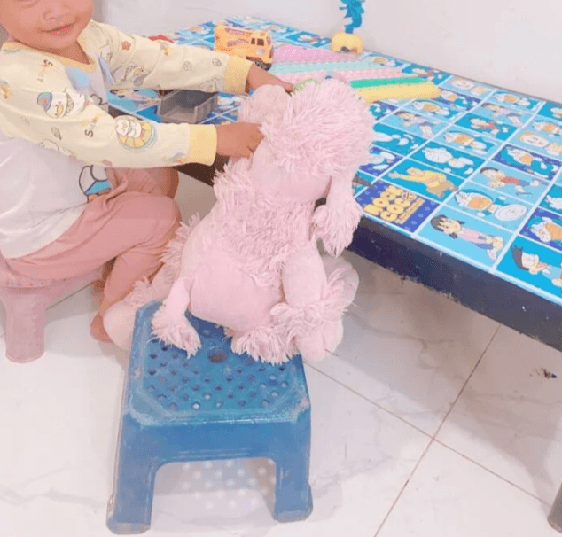 A toddler boy smiling as he shows a pink stuffed poodle his toy trucks on a Doraemon table.
