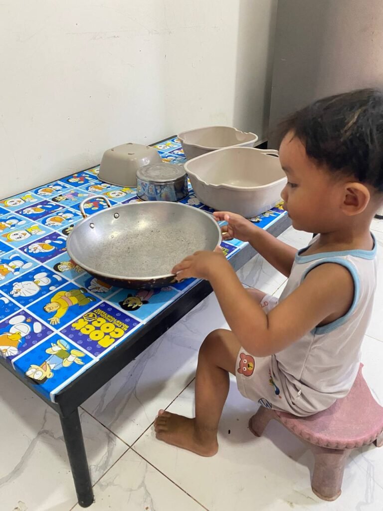 A toddler boy sitting at a blue Doraemon table looking at a metal frying pan and plastic basins for quiet play.