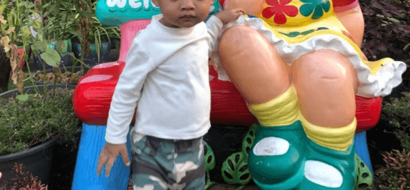 A toddler boy stands next to a brightly colored garden statue, looking serious and slightly upset about his outfit.