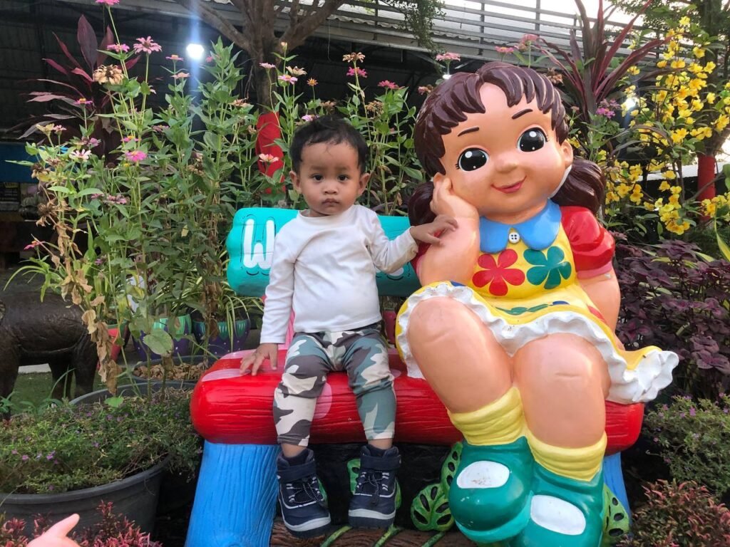 Toddler boy sitting on a colorful garden bench next to a smiling girl statue wearing a bright yellow flower dress.