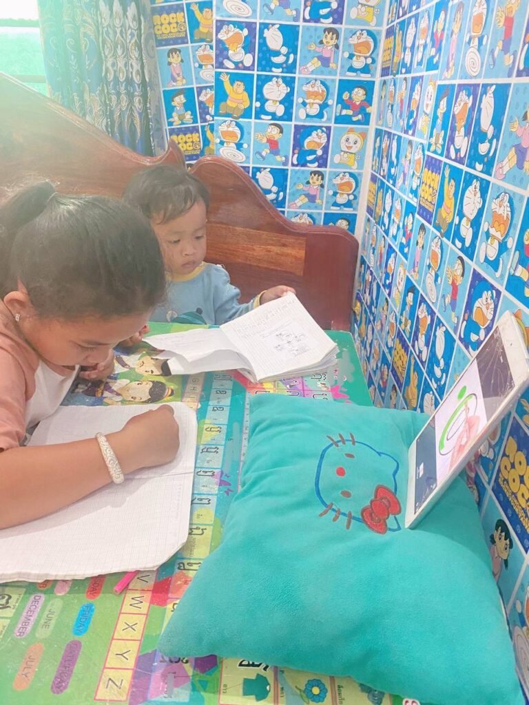 2-year-old boy watching intently as his 8-year-old sister reads and writes in her notebook.
