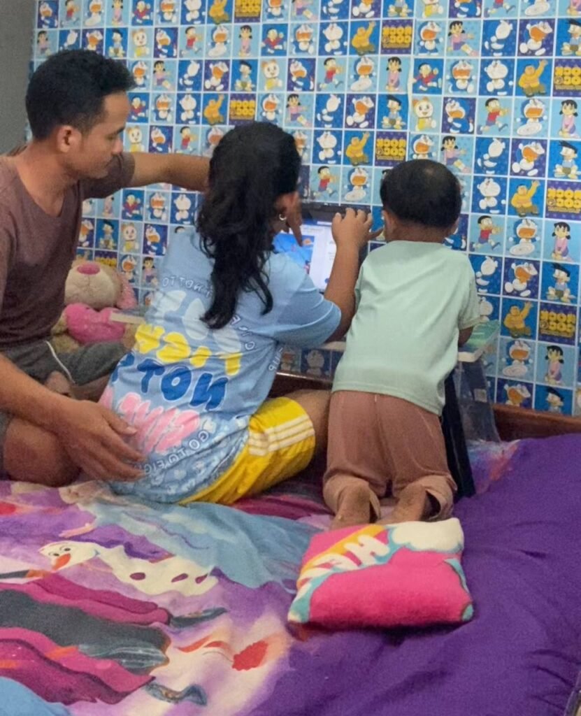 Toddler boy touching laptop screen to identify letters while father and sister watch, showing active engagement.